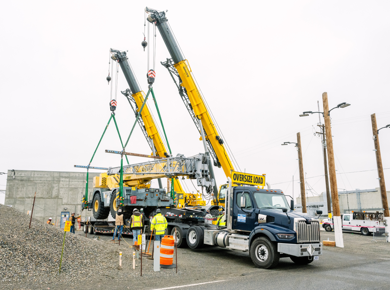 Two large cranes lower a 70-ton crane onto the back of a flatbed truck. 